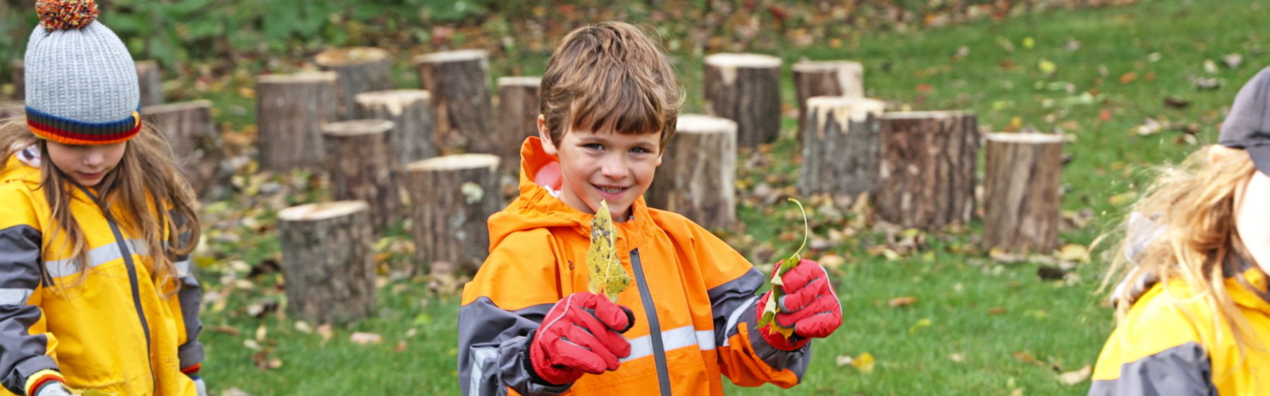 three outdoor kindergarten students, student in the middle is holding leaves
