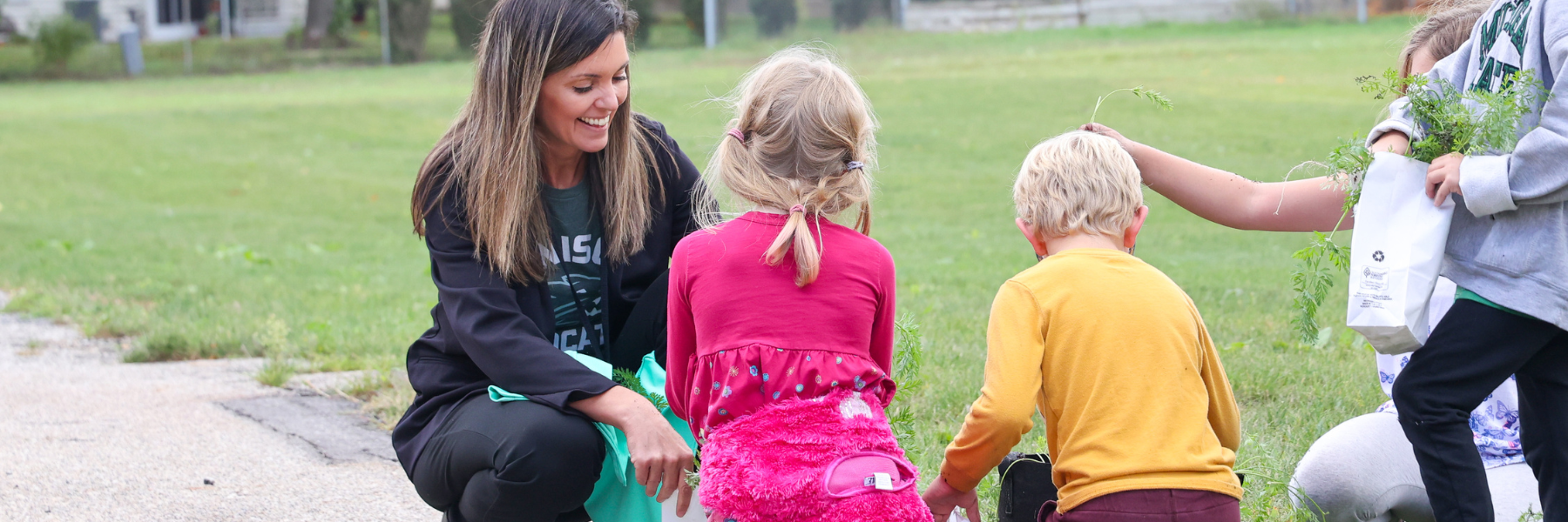 A teacher holds open a bag for students to place vegetables in outside.