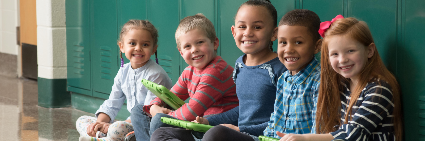 Elementary students sit against green lockers with iPads, smiling at the camera.
